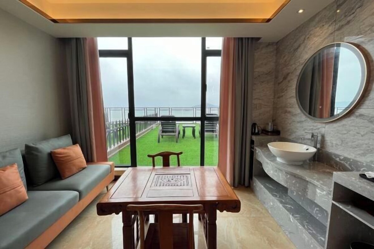 A modern hotel room with a large window, a sofa, a dining table, and a bathroom with a large mirror and sink.