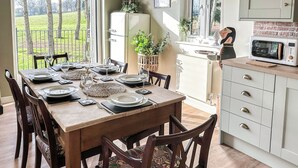 Un coin repas avec une table en bois dressée pour six personnes, entourée de chaises dotées de coussins à motifs. Un réfrigérateur blanc et un four à micro-ondes sont visibles à l’arrière-plan.