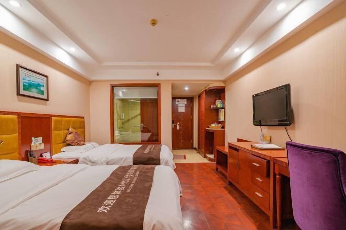 Habitación de hotel con dos camas, un escritorio, un televisor y un baño visible a través de una puerta de vidrio.