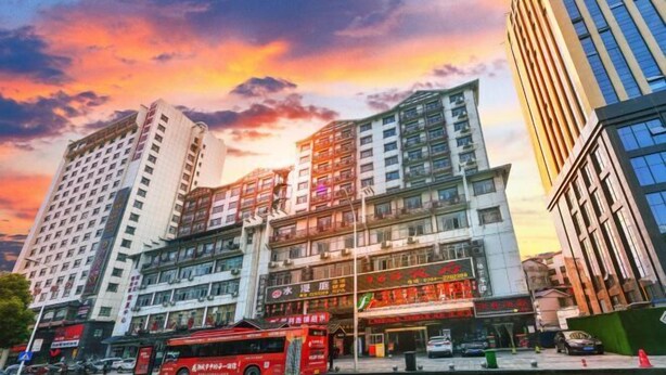 A multi-story building with commercial spaces on the ground floor, a red bus in front, and a sunset sky. - Fulante Le Hotel (Tianmen Mountain Fengwan Road) (Zhangjiajie)