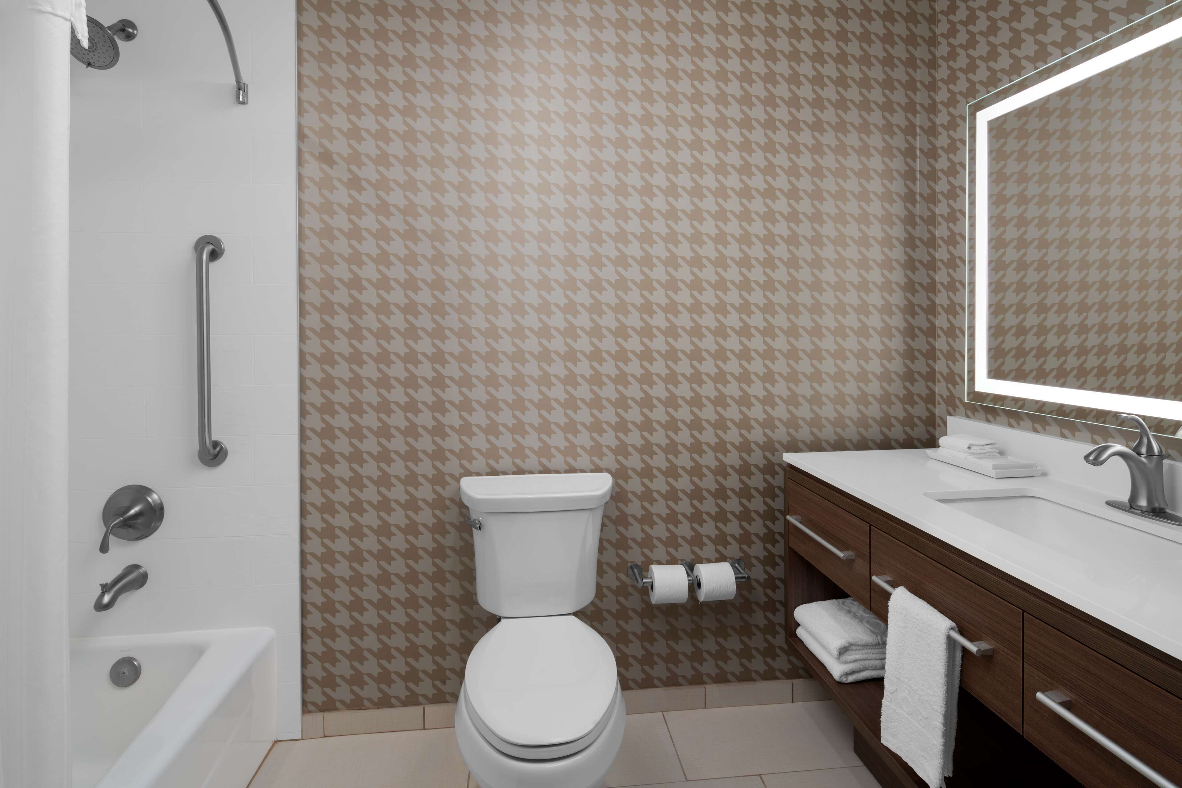 Un baño con inodoro blanco, bañera, ducha con barra de apoyo, lavamanos con espejo y azulejos con diseño.