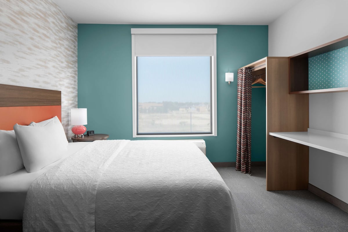 A hotel room with a bed, a nightstand, a lamp, a window with a view, a wardrobe, and a shelf.