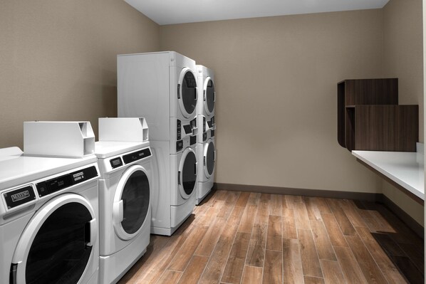 A laundry room with two front-loading washing machines, a dryer, and a wooden floor. - Home2 Suites By Hilton Hobbs (Hobbs)