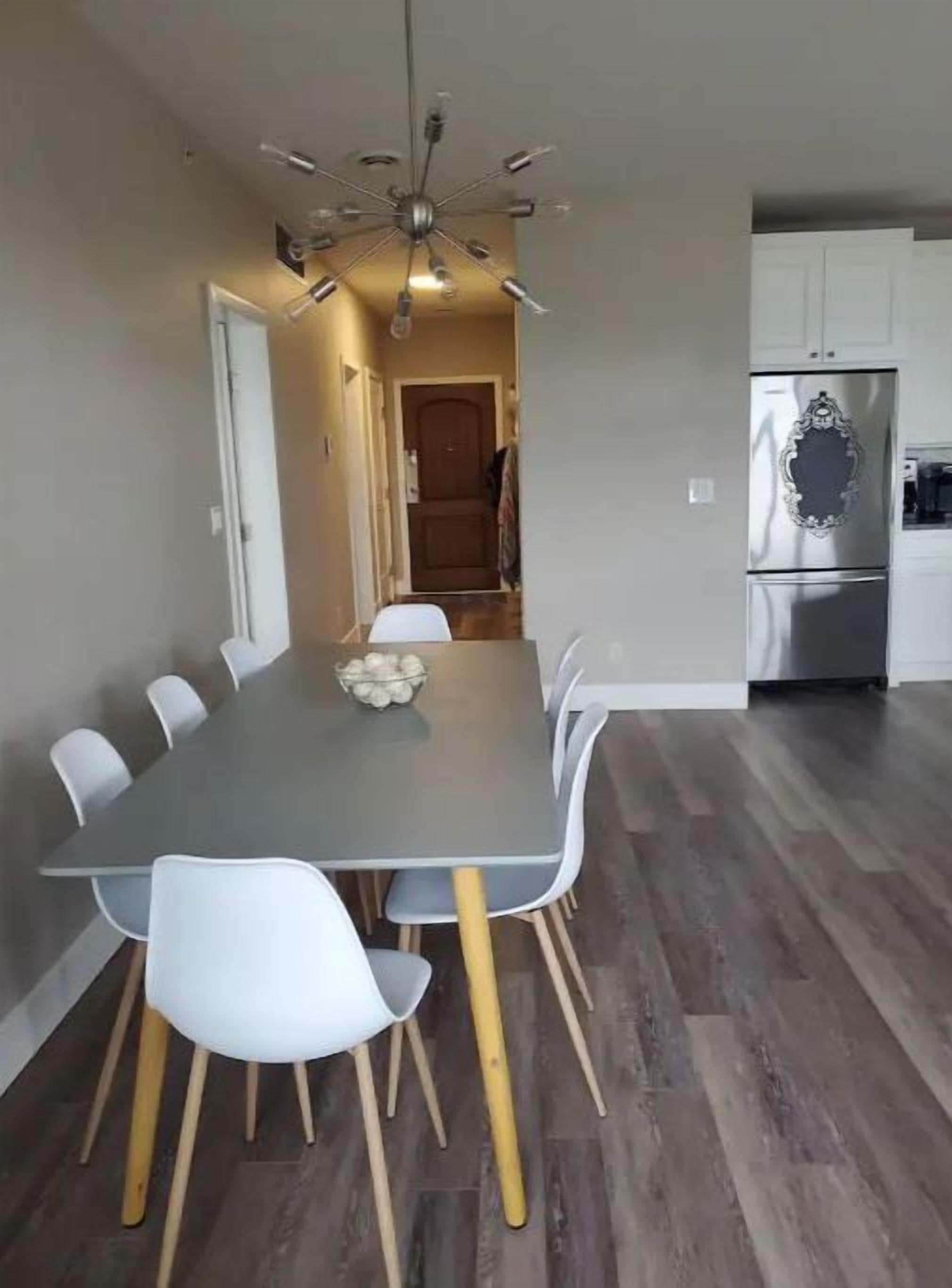 Une cuisine moderne avec un réfrigérateur en acier inoxydable, un coin repas doté d’une table grise et de chaises blanches, et un plancher en bois.