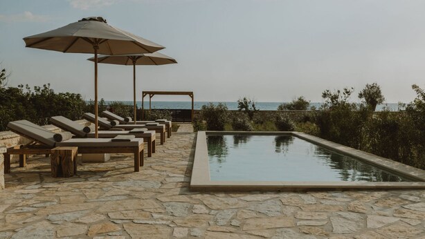 A poolside area with lounge chairs and umbrellas, a stone-tiled floor, and a view of the sea. - Nama Seaside Villas (Preveza)