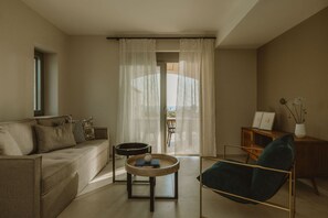 A living room with a sofa, a round coffee table, a chair, and a view through a window with curtains. - Nama Seaside Villas (Preveza)