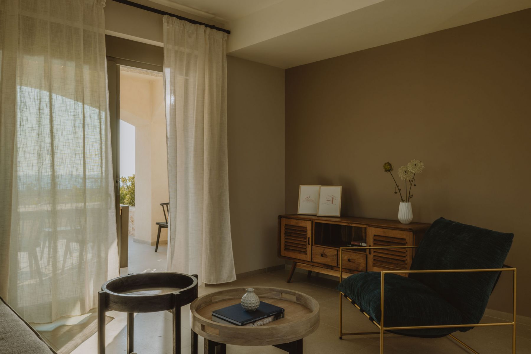 Una habitación con vistas a través de una ventana, un armario de madera, una silla azul y una mesita con un libro y un jarrón.
