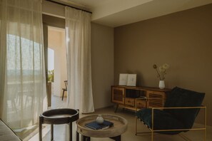A room with a view through a window, a wooden cabinet, a blue chair, and a small table with a book and a vase. - Nama Seaside Villas (Preveza)