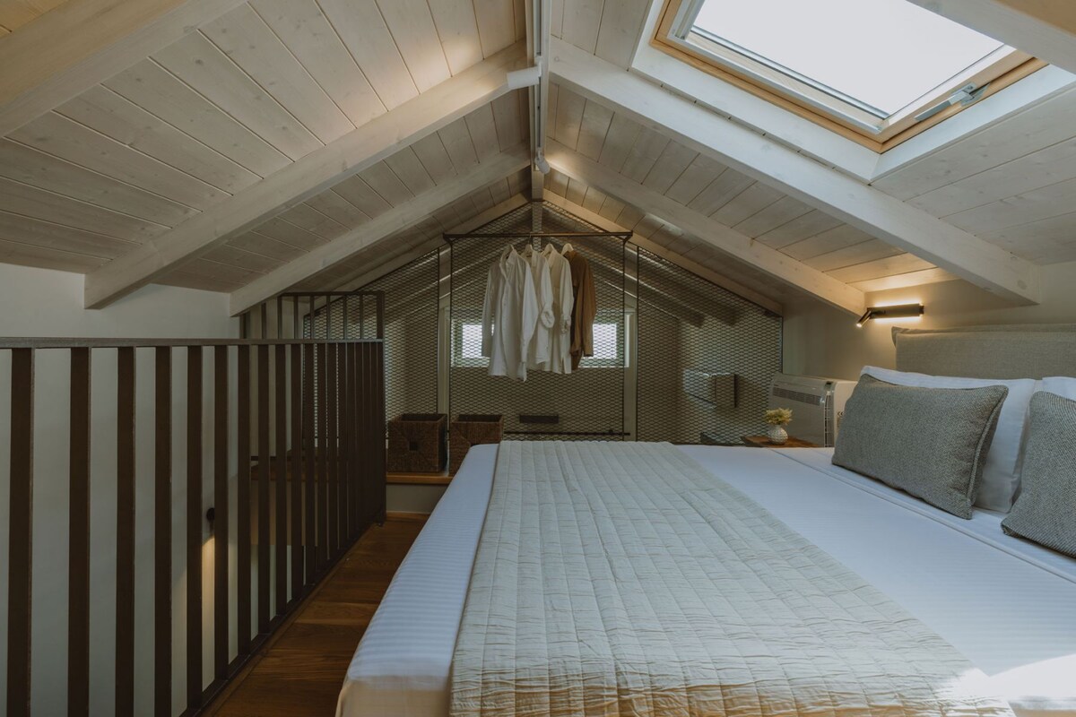 A bedroom with a sloped ceiling, a bed, a small table with a lamp, and a window.