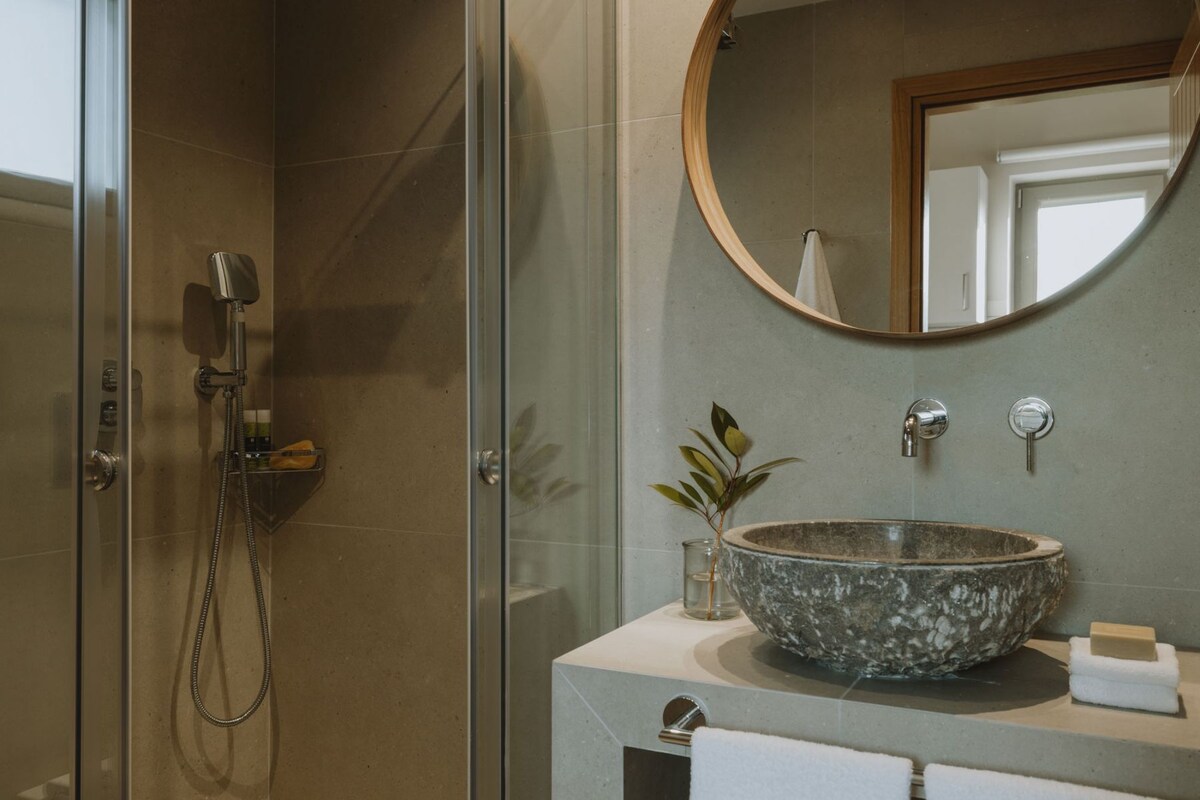 A bathroom with a glass-enclosed shower, a round mirror, a stone sink, and a small plant on the countertop.