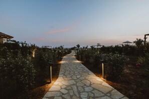 A stone pathway leading through a garden with plants on either side, flanked by structures with umbrellas. - Nama Seaside Villas (Preveza)