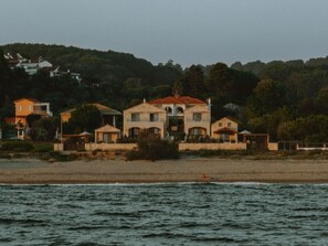 Una propiedad costera con varias construcciones, una playa al frente y una colina boscosa al fondo.