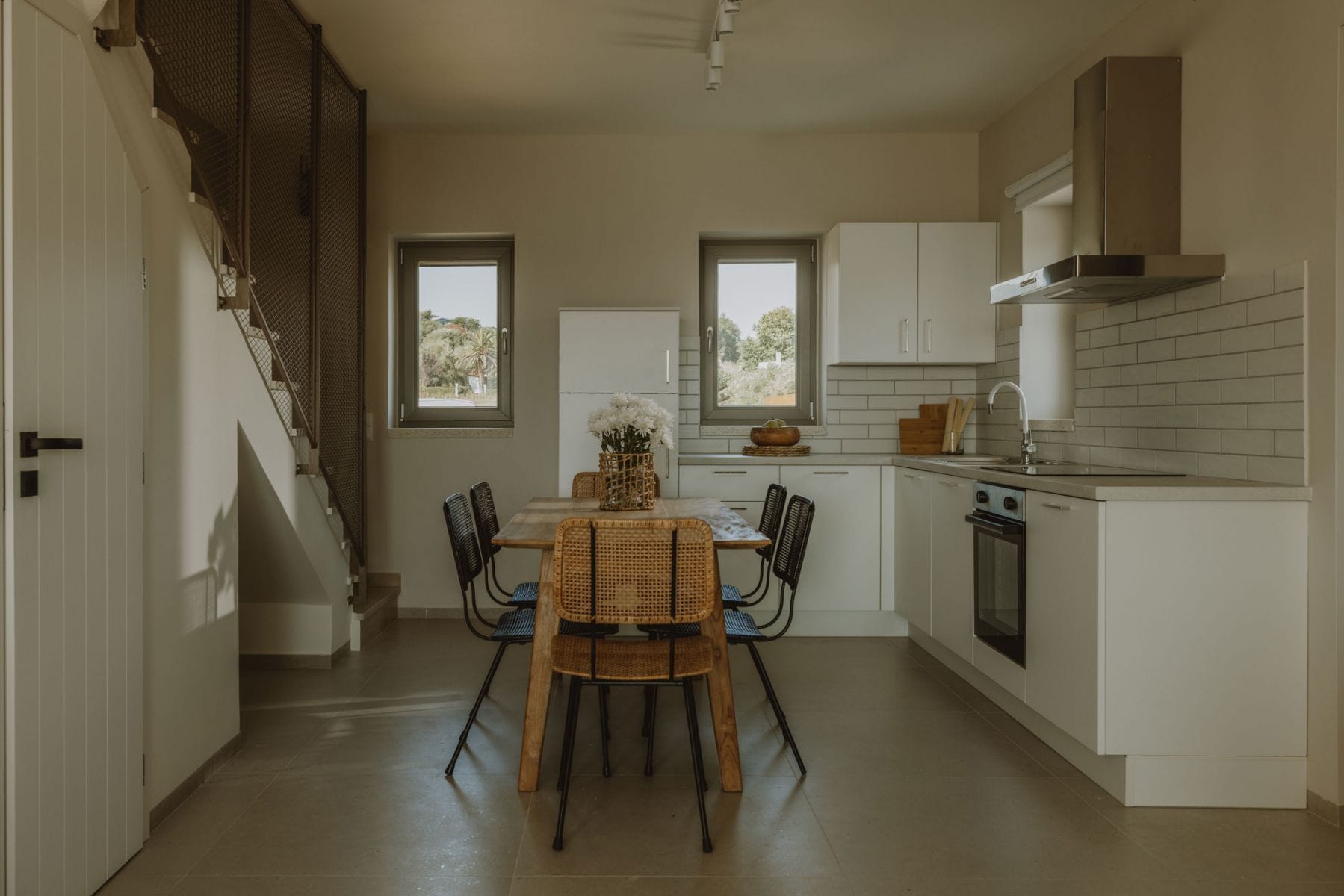 A modern kitchen with white cabinetry, a dining area with wicker chairs, and a staircase on the left.