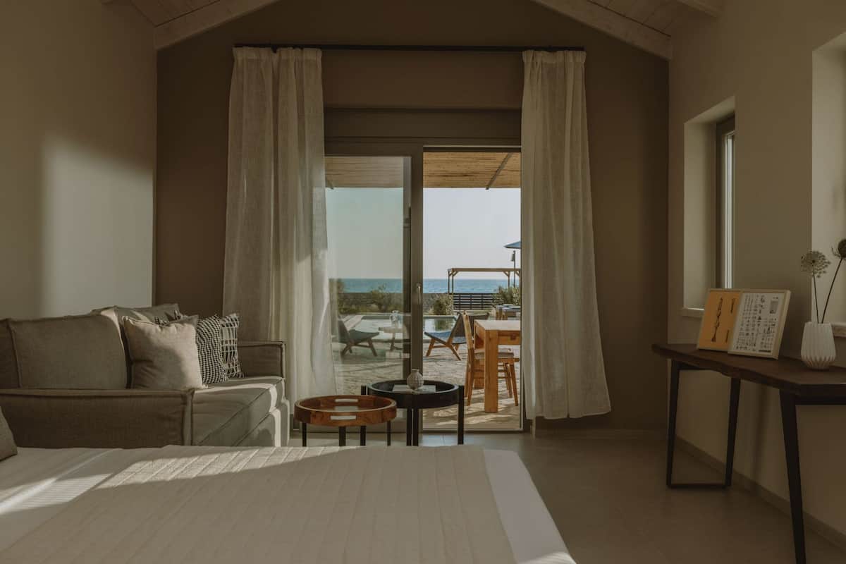 A bedroom with a bed, a sofa, a table, and a view of the outdoors.