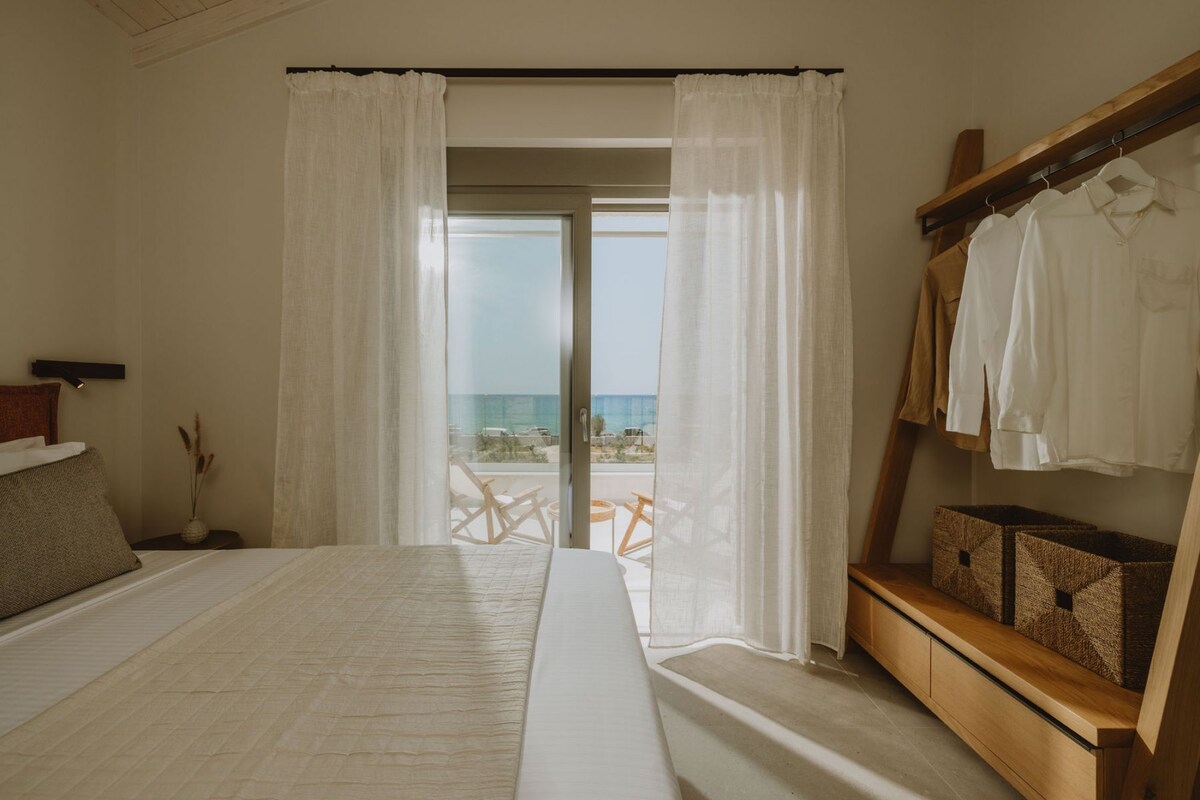 A bedroom with a large bed, a wooden bench with storage, a view of the beach, and a balcony with a table and chairs.