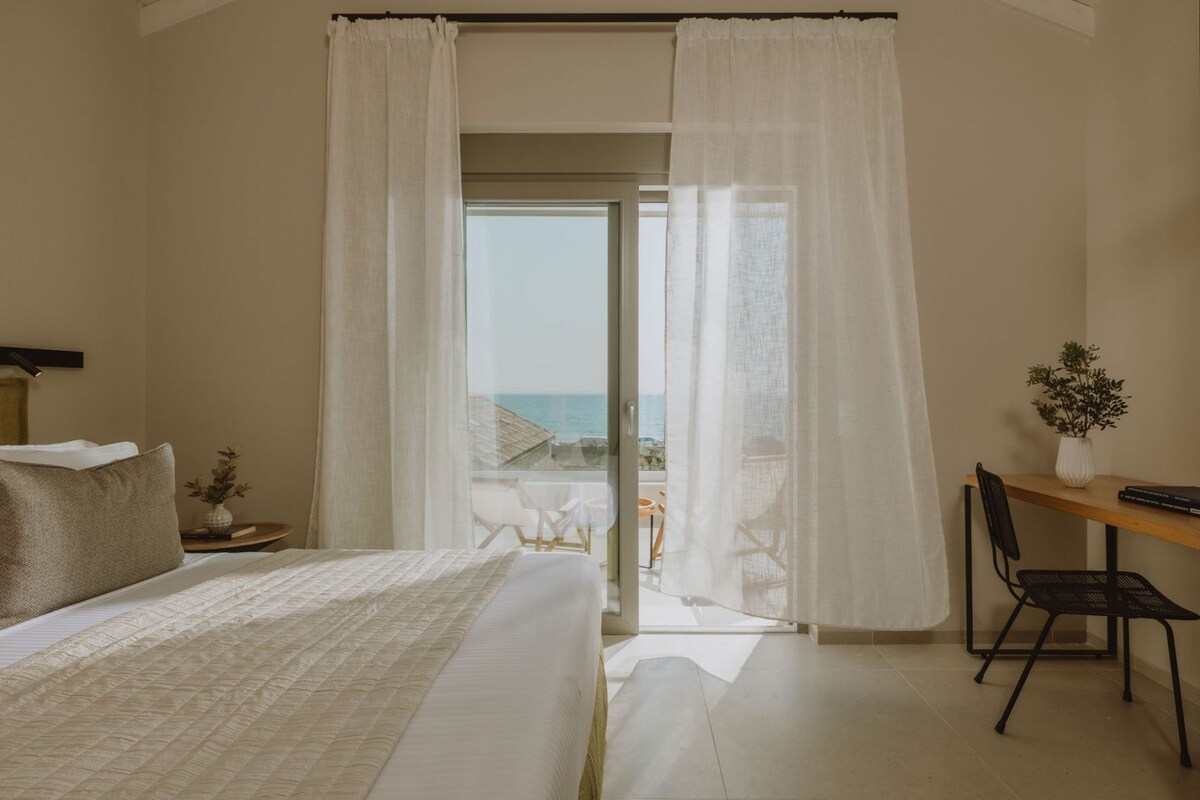 A bedroom with a bed, a desk, and a view of the sea.
