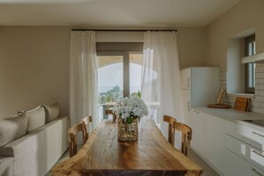 Un comedor con mesa de madera, sillas, un jarrón con flores y vistas a través de una ventana con cortinas.