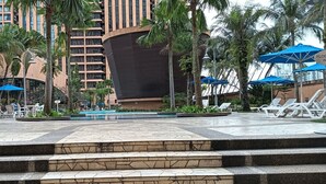 Area piscina di un hotel con lettini e ombrelloni, circondata da palme e un edificio dalla facciata curva.