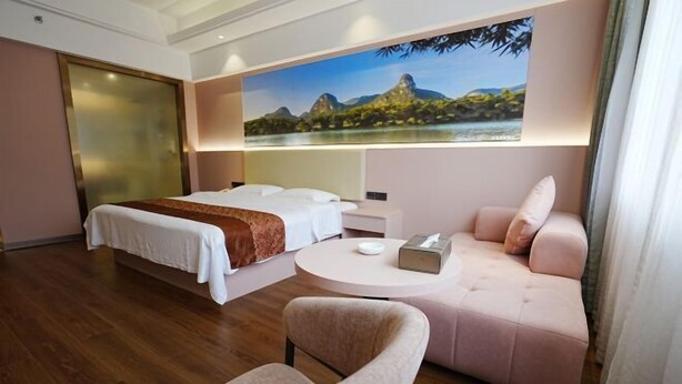 A hotel room with a bed, a sofa, a chair, a round table, and a painting of a landscape on the wall. - Ningxin International Hotel (Luocheng)