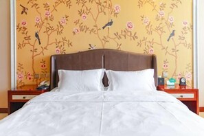 A neatly made bed with white linens, a wooden headboard, and a floral wallpaper design on the wall behind.