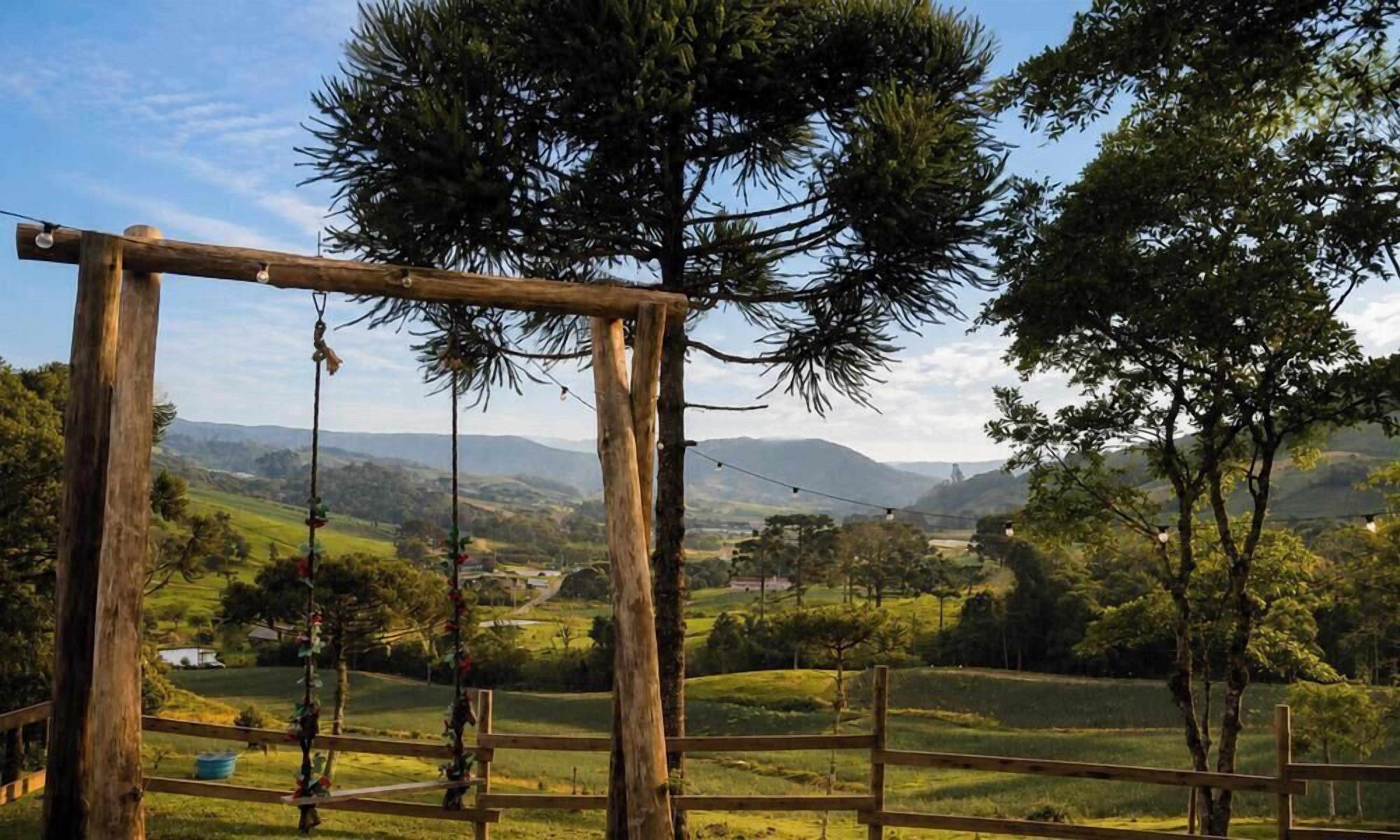 Une balançoire en bois, munie de cordes, entourée d’une clôture en bois, et offrant une vue sur un paysage verdoyant avec des arbres et des collines lointaines.