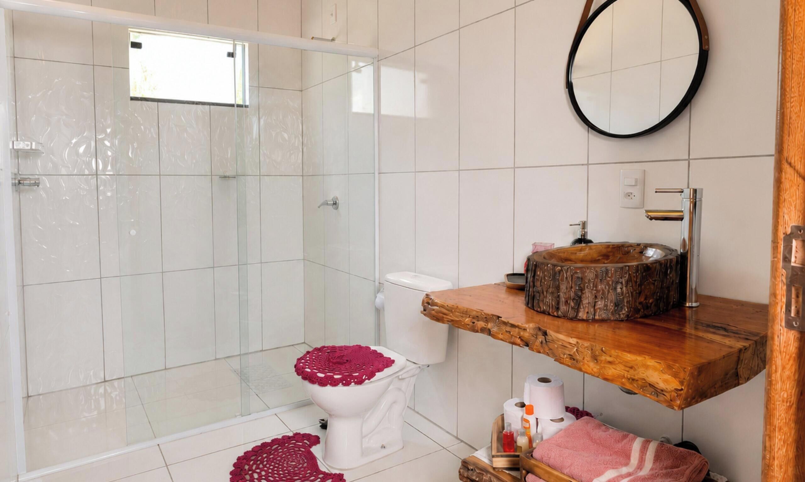 Un baño moderno con cabina de ducha de vidrio, un inodoro blanco con dispensador rojo de papel higiénico, un mueble de tocador de madera con un lavabo redondo y un espejo en la pared.
