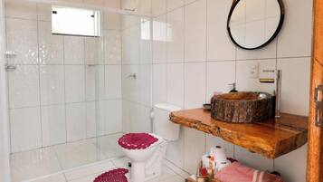 Un baño moderno con cabina de ducha de vidrio, un inodoro blanco con dispensador rojo de papel higiénico, un mueble de tocador de madera con un lavabo redondo y un espejo en la pared.