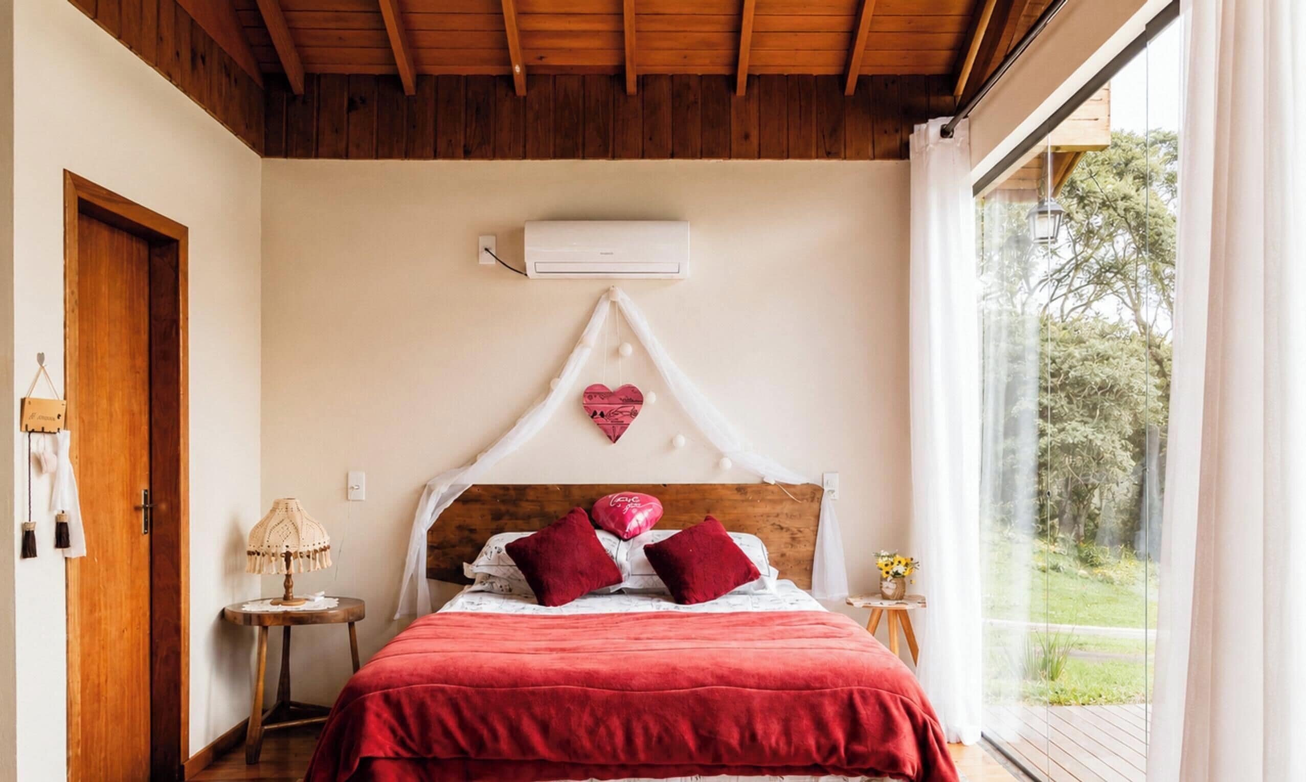 Ein Schlafzimmer mit Holzbalkendecke, einem Bett mit roter Bettwäsche, einer herzförmigen Dekoration und einem Fenster mit Blick auf Grünflächen.
