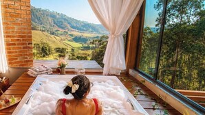 A person relaxing in a hot tub with a scenic view of hills and trees. - Chalé Caminho do Sol (Rancho Queimado)
