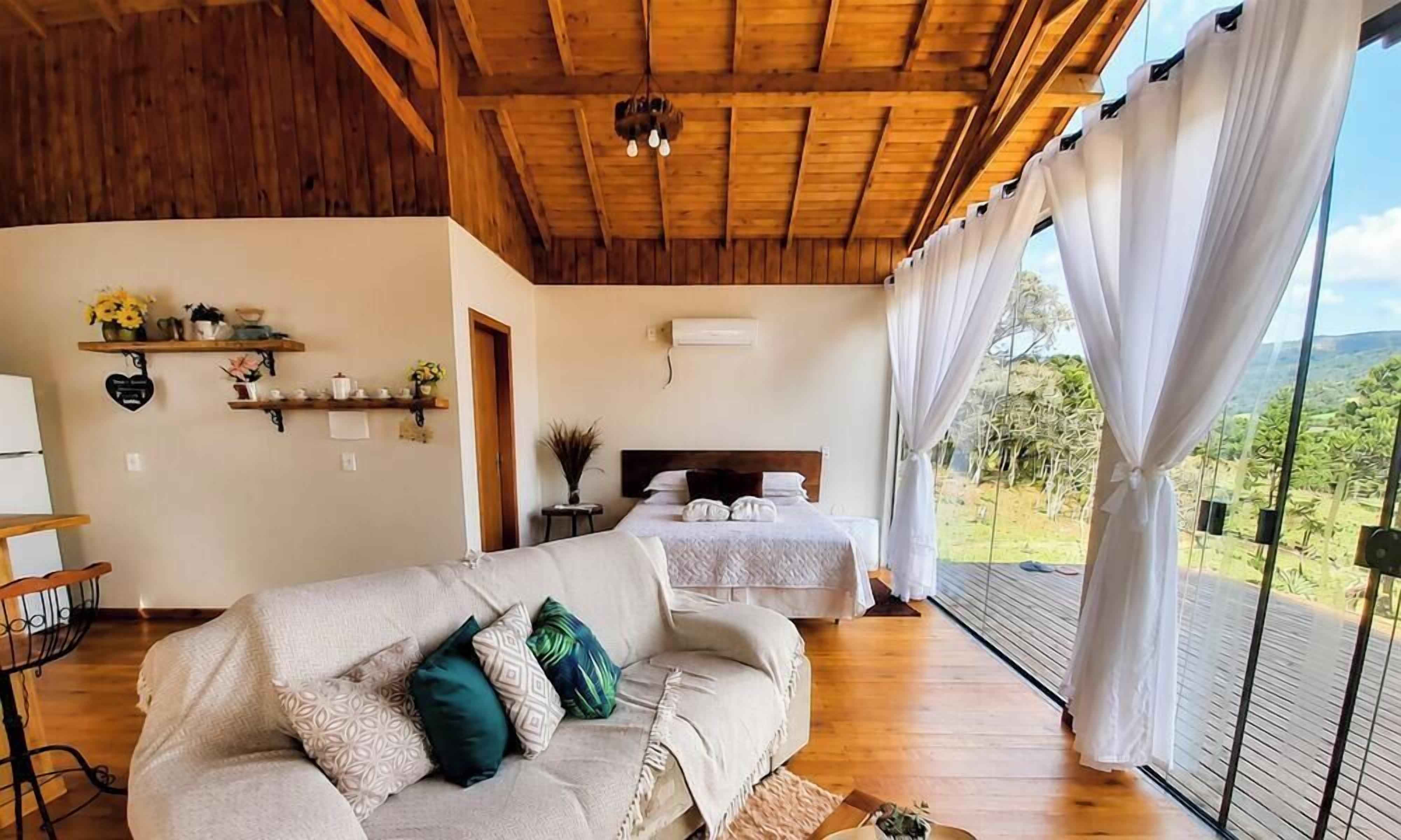 A room with a wooden ceiling, a sofa, a bed, a window with curtains, and a view of the outdoors.