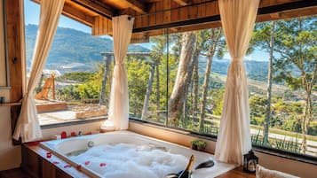 A spa room with a large bathtub filled with bubbles, a bottle of champagne, and a view of a mountainous landscape through large windows.