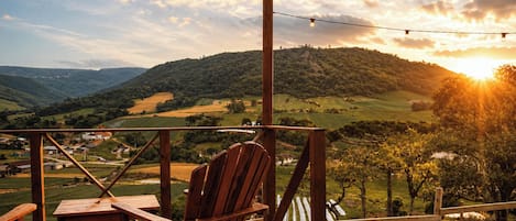 A wooden deck with a chair and table, overlooking a hilly landscape with a sunset.