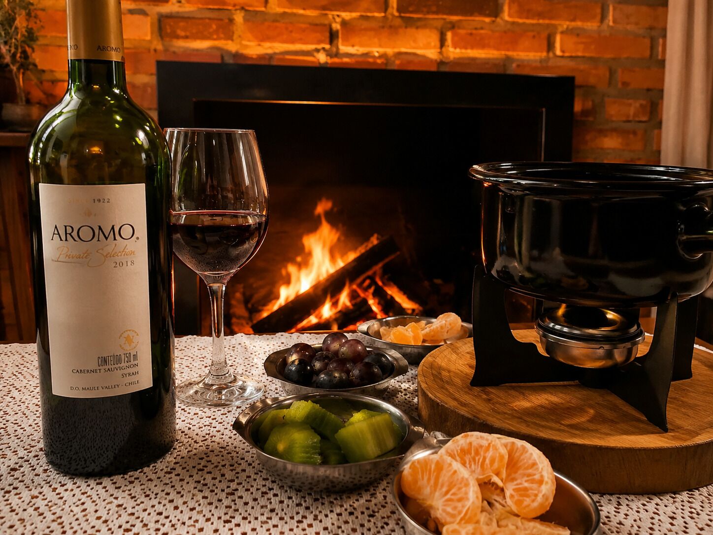 A bottle of AROMO Private Selection 2018 wine, a glass of wine, a bowl of fruits, and a black pot on a wooden board, all set in front of a fireplace.