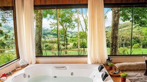 A room with a large window, a bathtub, and a view of a green landscape. - Chalé Caminho do Sol (Rancho Queimado)