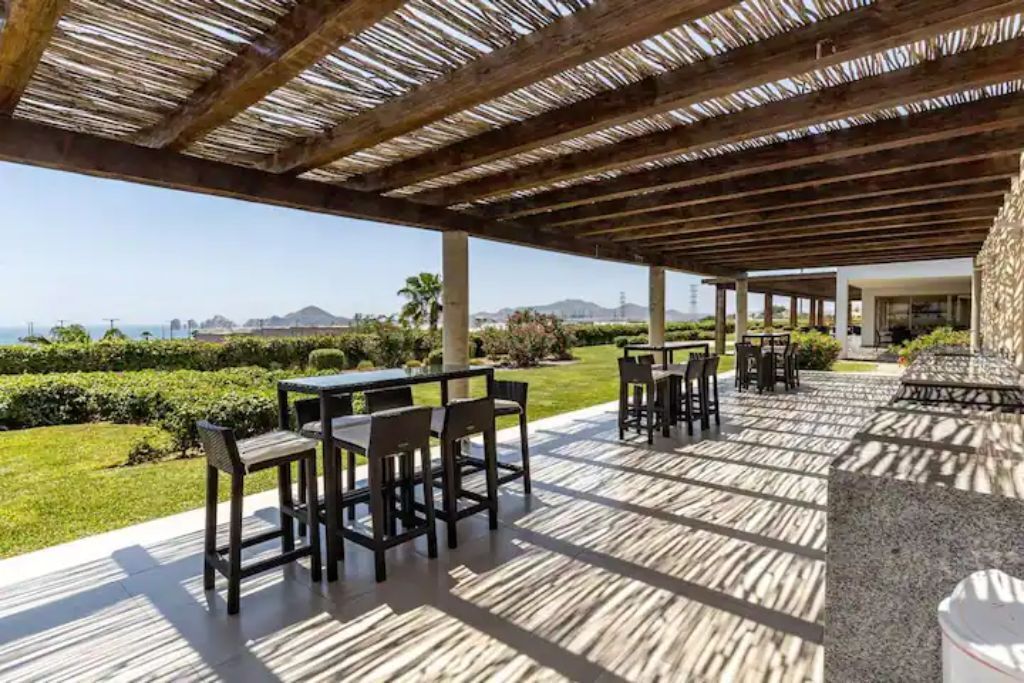 An outdoor patio with tables and chairs, a stone wall, and a view of the ocean and hills.