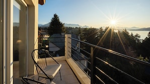 En balkon med en stol, glasdør og udsigt til bjerge og sø ved solnedgang.