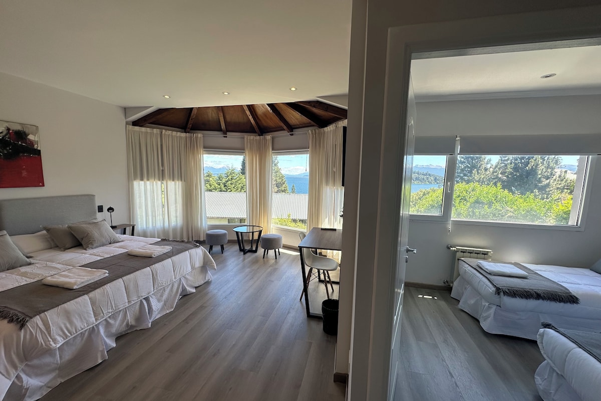 A modern hotel room with a large bed, a smaller bed, a dining area, and a view of the outdoors.