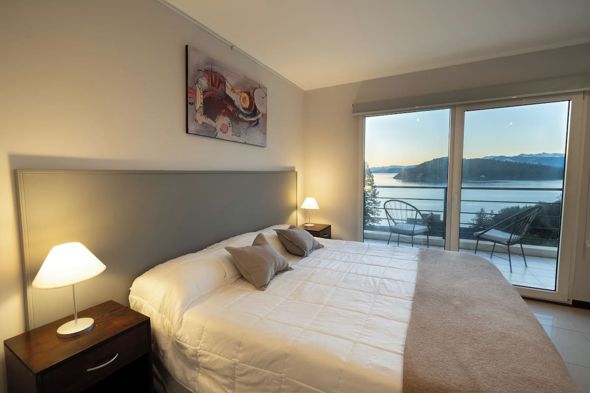 Ein Schlafzimmer mit einem großen Bett, zwei Nachttischlampen, einem Gemälde an der Wand und Seeblick durch eine Schiebetür.