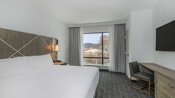 Une chambre d’hôtel avec un grand lit, un bureau, une chaise, une télévision et une fenêtre donnant sur des immeubles et des collines.