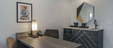A modern hotel room with a wooden dining table, a single chair, a desk with a lamp, a microwave, and a large mirror.