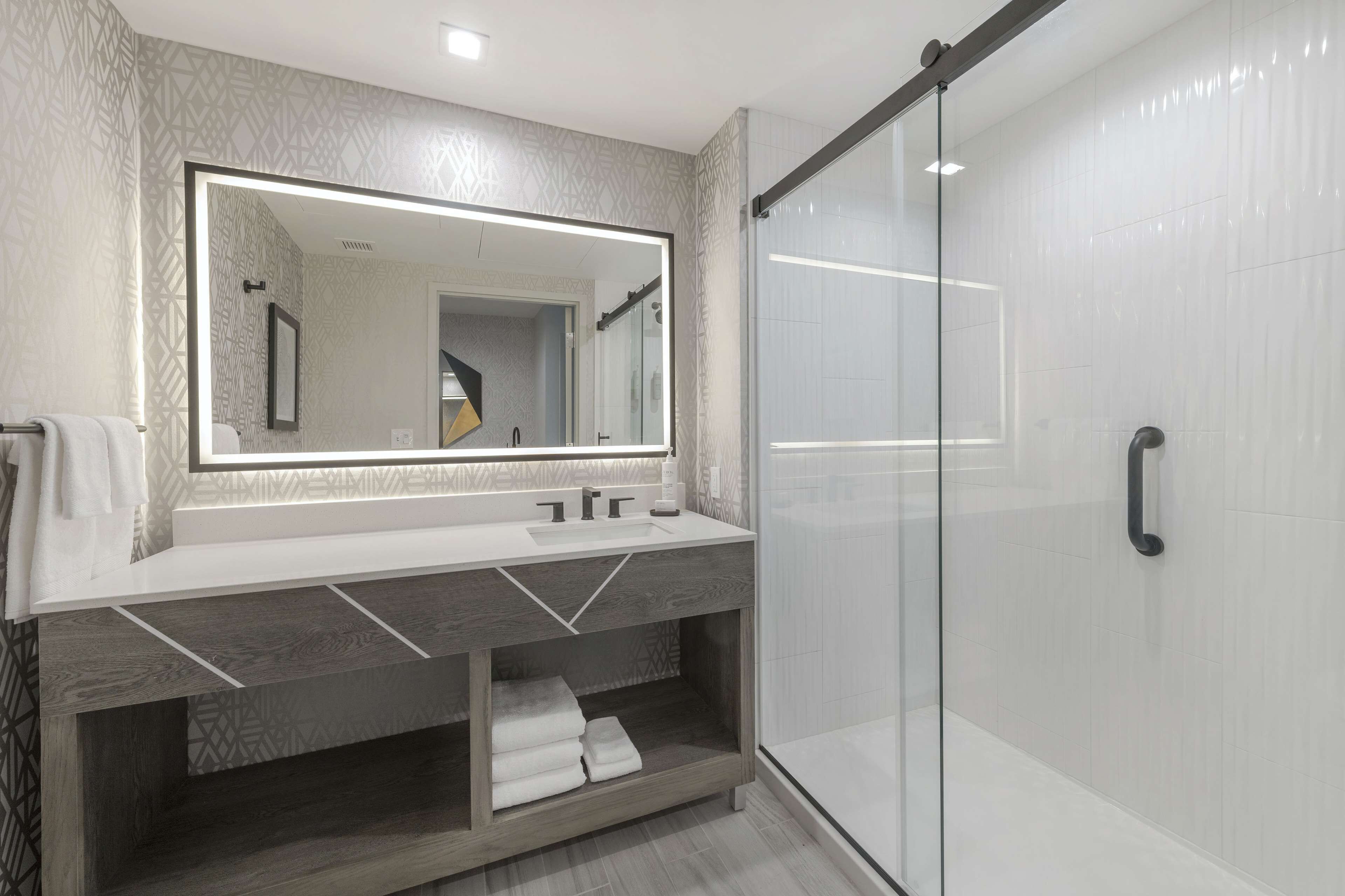 A modern bathroom with a large mirror, a glass shower enclosure, and a vanity with a towel rack.
