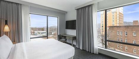 A hotel room with a large bed, a desk, and a TV. The room has large windows with a view of buildings and a cityscape.