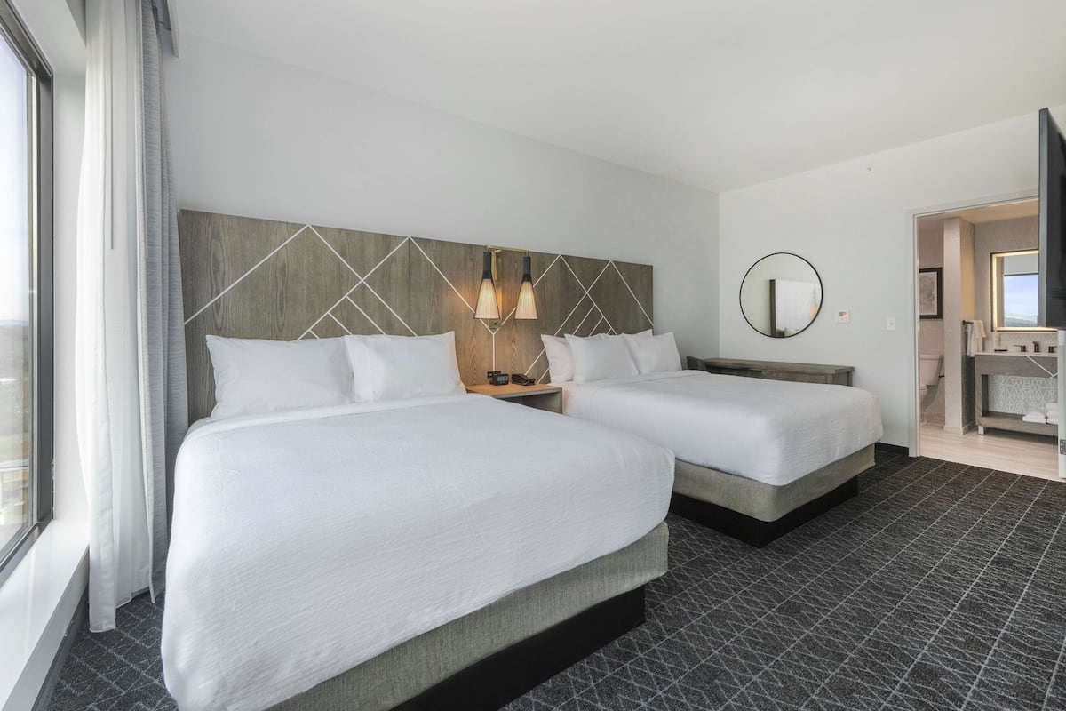 A hotel room with two beds, a large window with curtains, a headboard with geometric patterns, a round mirror, and a small bedside table with a lamp.