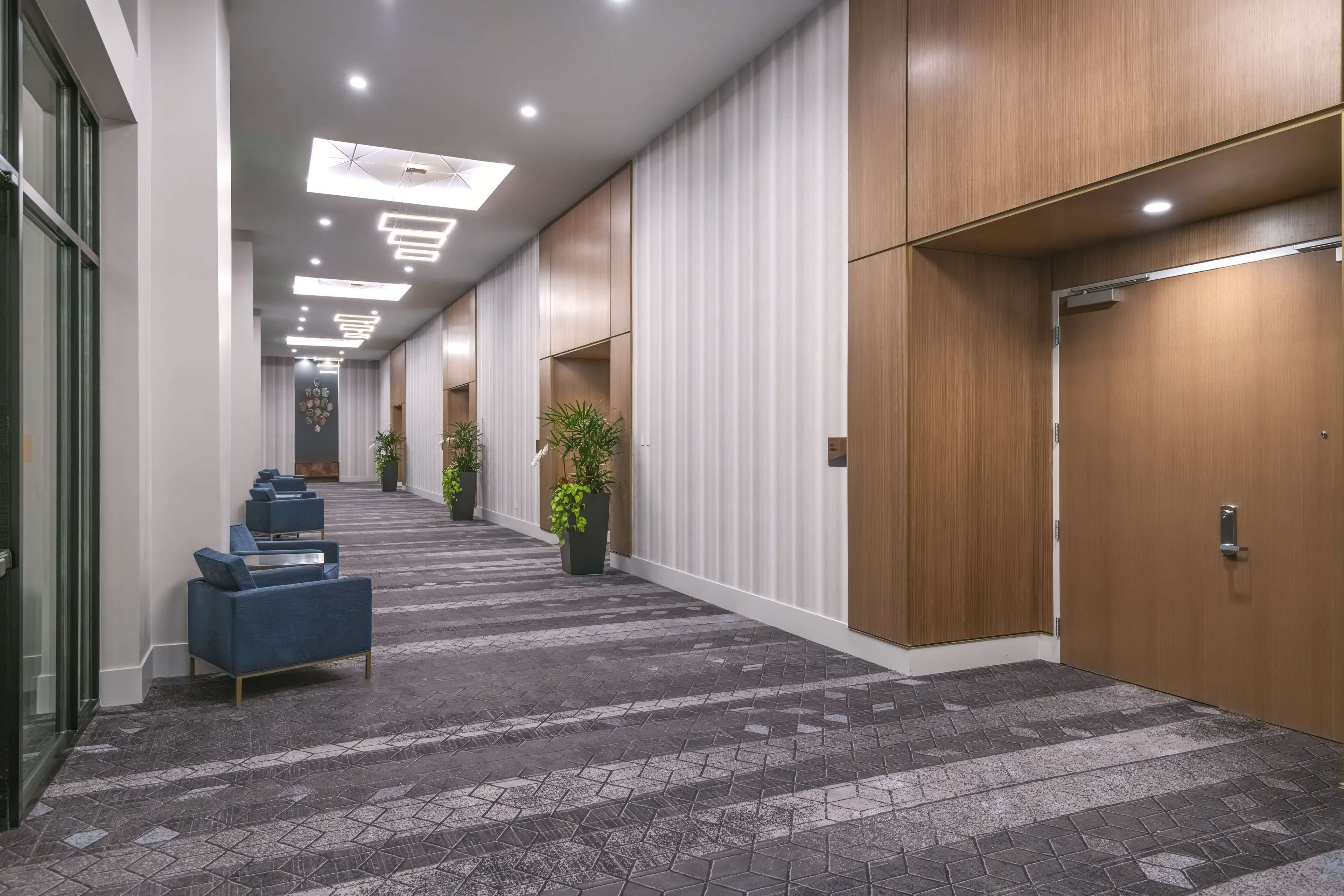 Corredor de hotel com carpete estampado, assentos azuis, plantas em vasos e portas de madeira.