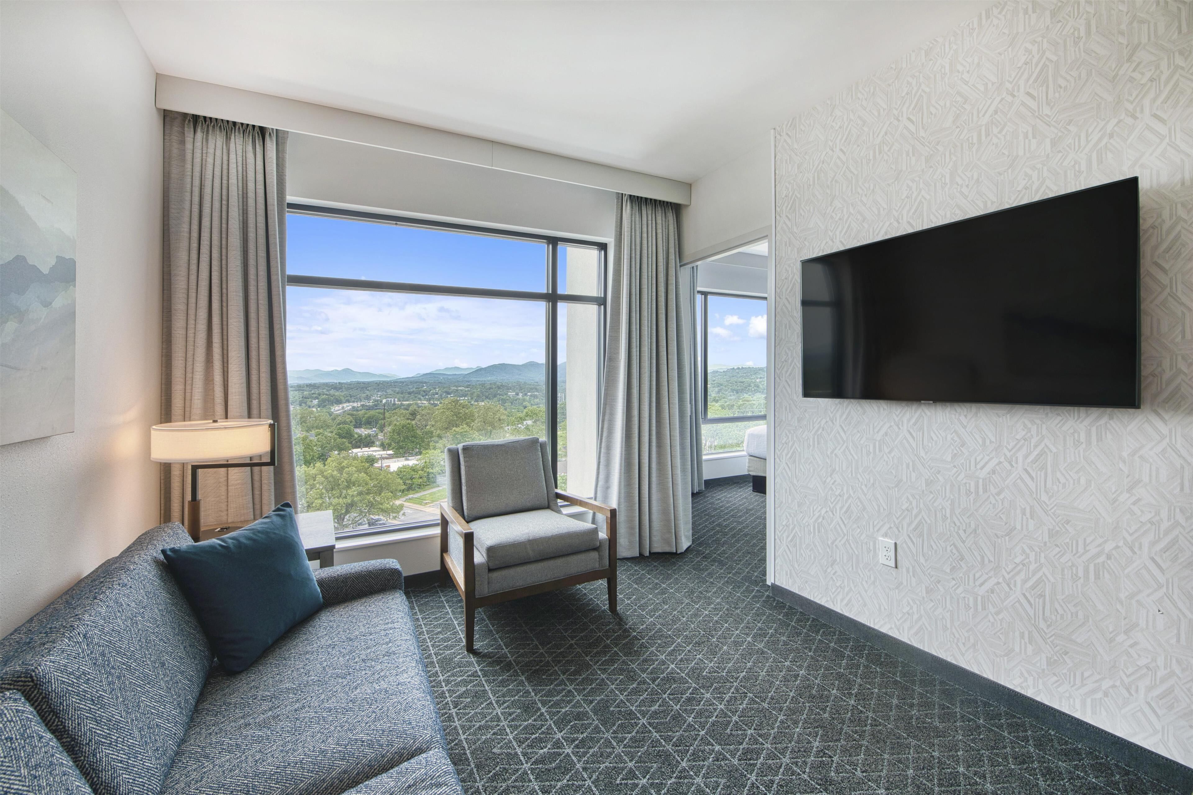 Una habitación de hotel con un ventanal grande, un televisor de pantalla plana, un sofá y una silla.