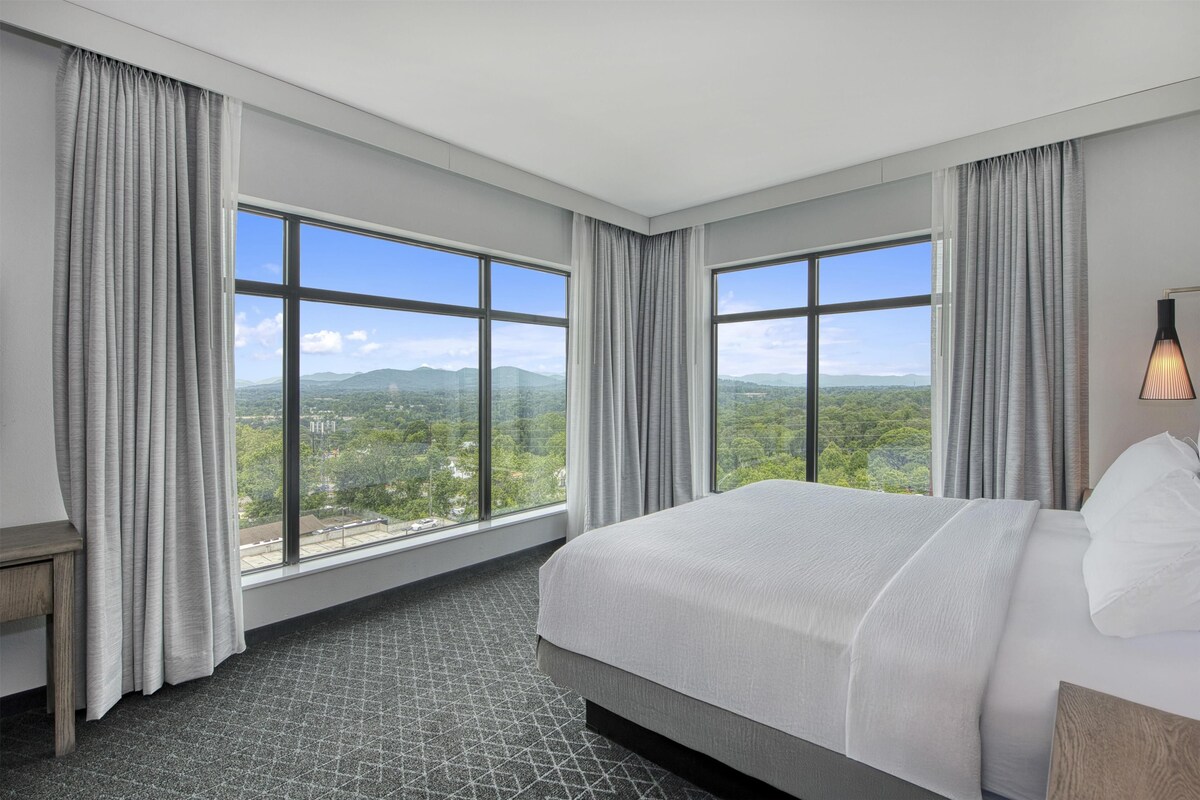 A hotel room with a large bed, two windows with curtains, a view of greenery, and a lamp on the nightstand.