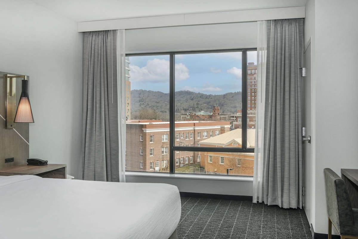 A hotel room with a large window, a bed, a desk with a lamp, and a chair.