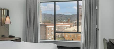 A hotel room with a large window, a bed, a desk with a lamp, and a chair.