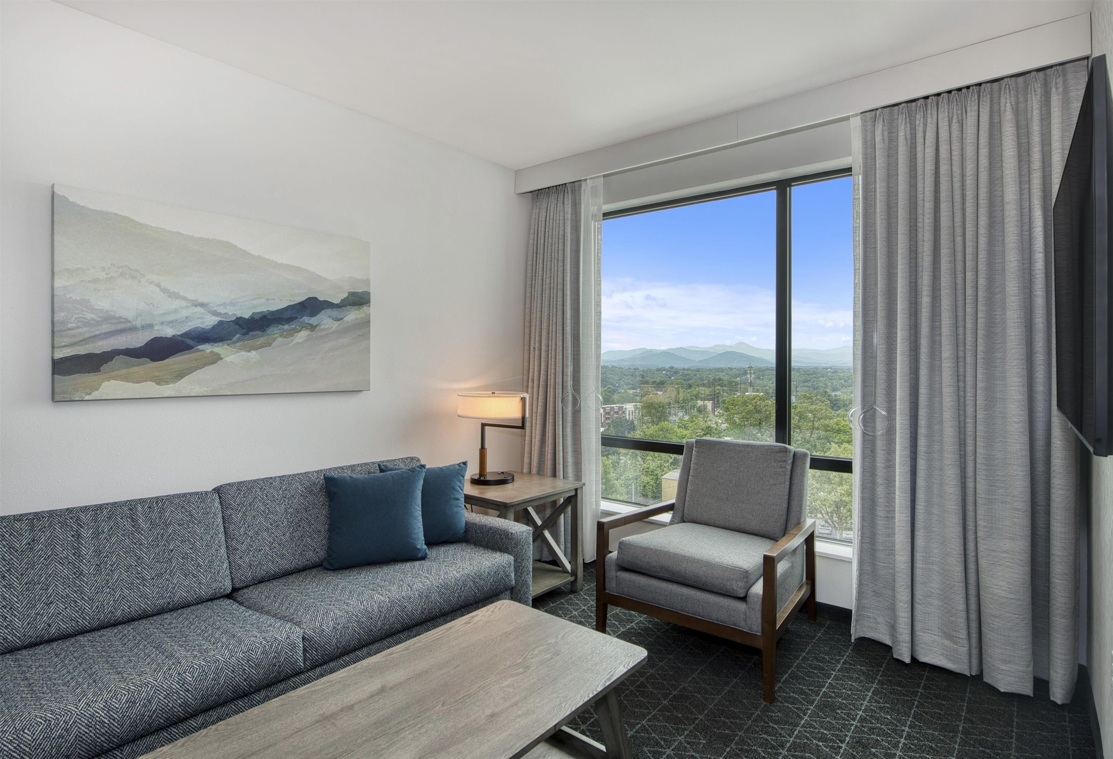 Ein Hotelzimmer mit Sofa, Sessel, Tisch, Lampe und einem großen Fenster mit Vorhängen.