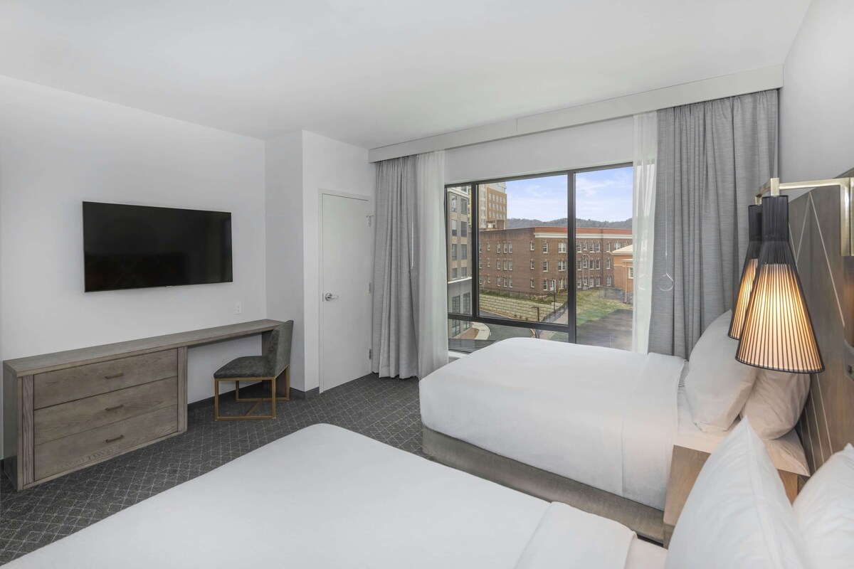 A hotel room with two beds, a TV mounted on the wall, a desk with drawers, a chair, and a large window with curtains.